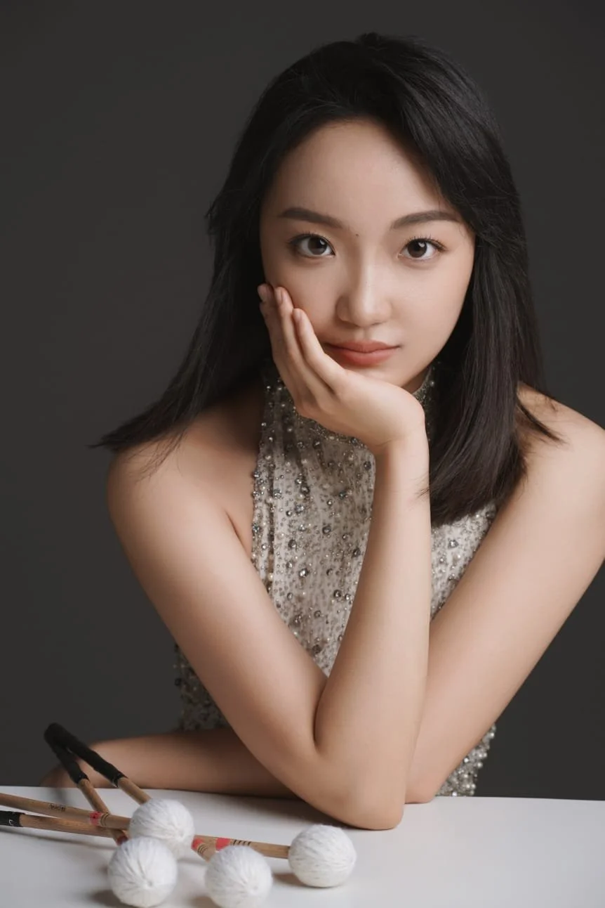 Woman posing with chin resting on hand, wearing a sequined dress, with percussion mallets on the table.