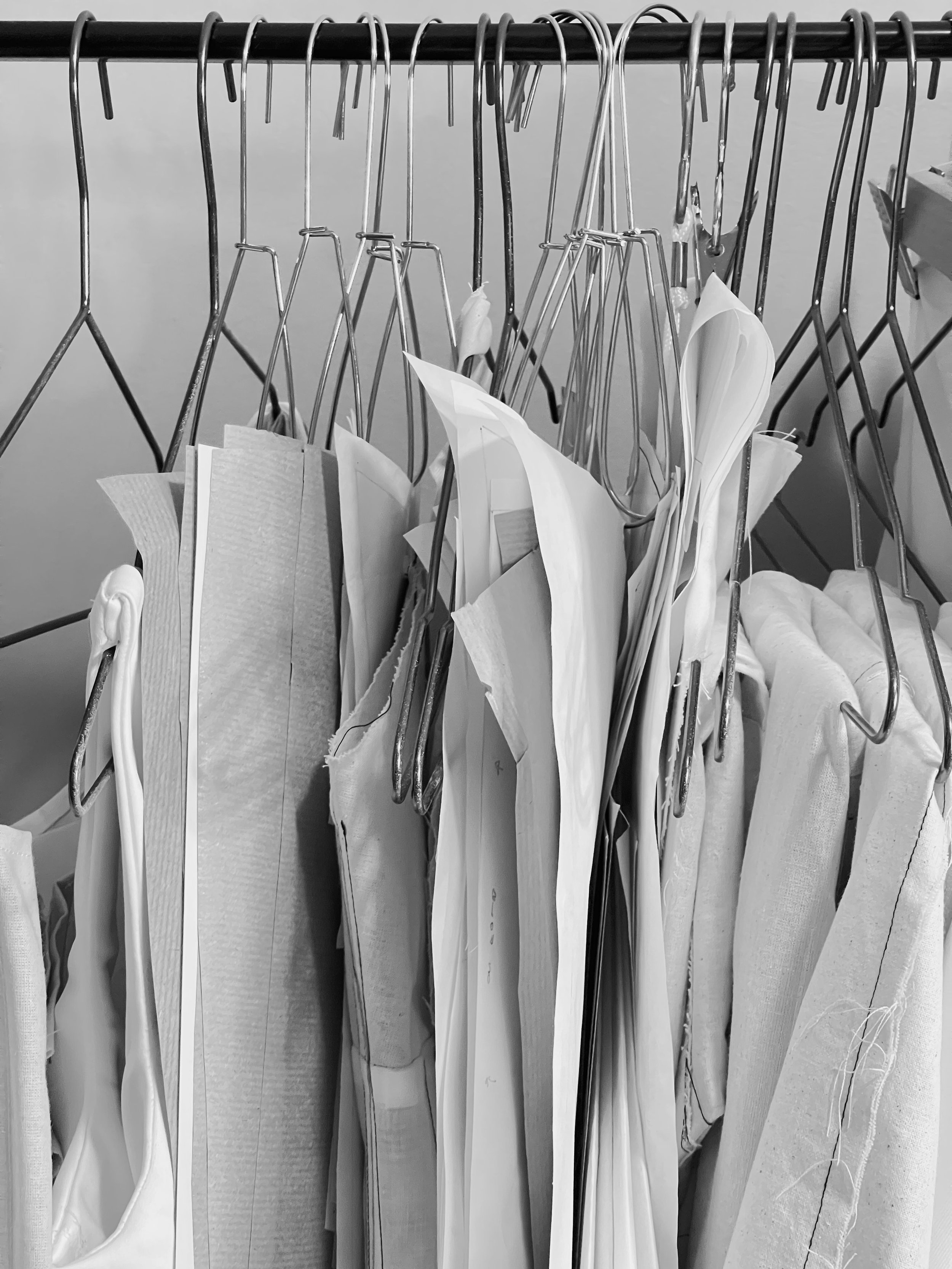 Black and white image of sewing patterns and fabric pieces hanging on metal hangers on a clothing rack.