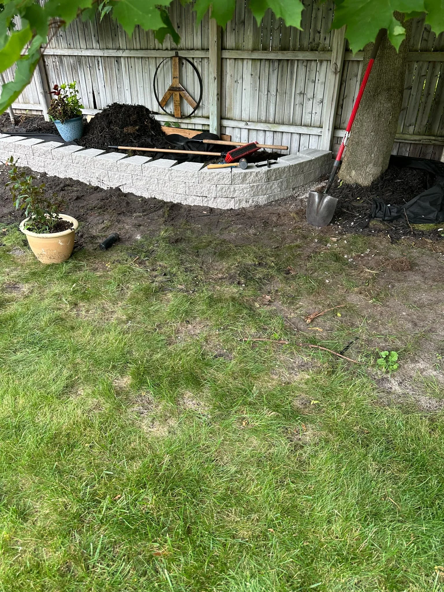 Garden area under construction with raised stone bed, mulch, a potted plant, tools, and a wooden fence.