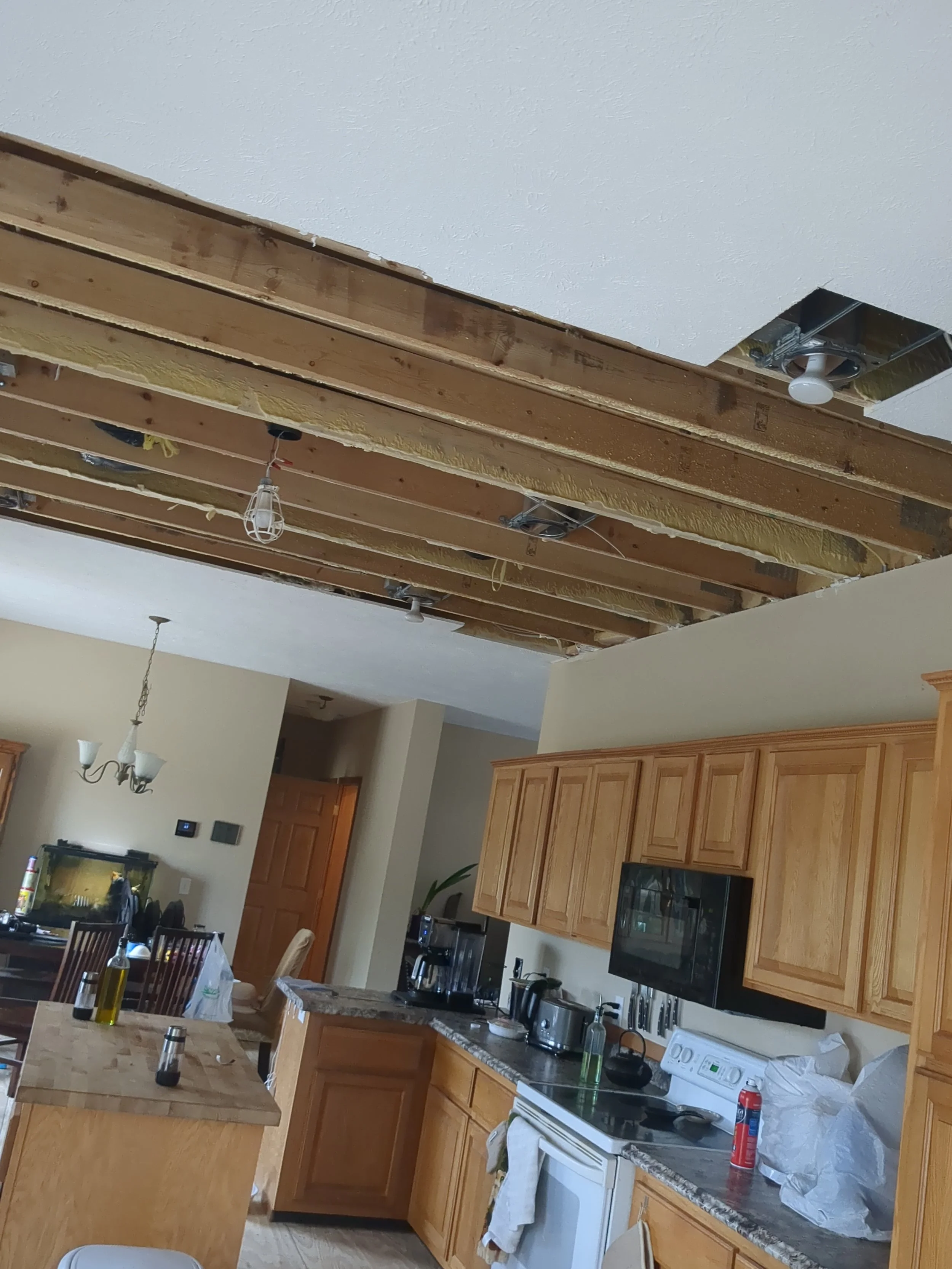 Kitchen with partially opened ceiling showing wooden beams, electrical wiring, and light fixtures. Wooden cabinets, countertops with kitchen appliances, and a dining area in the background.