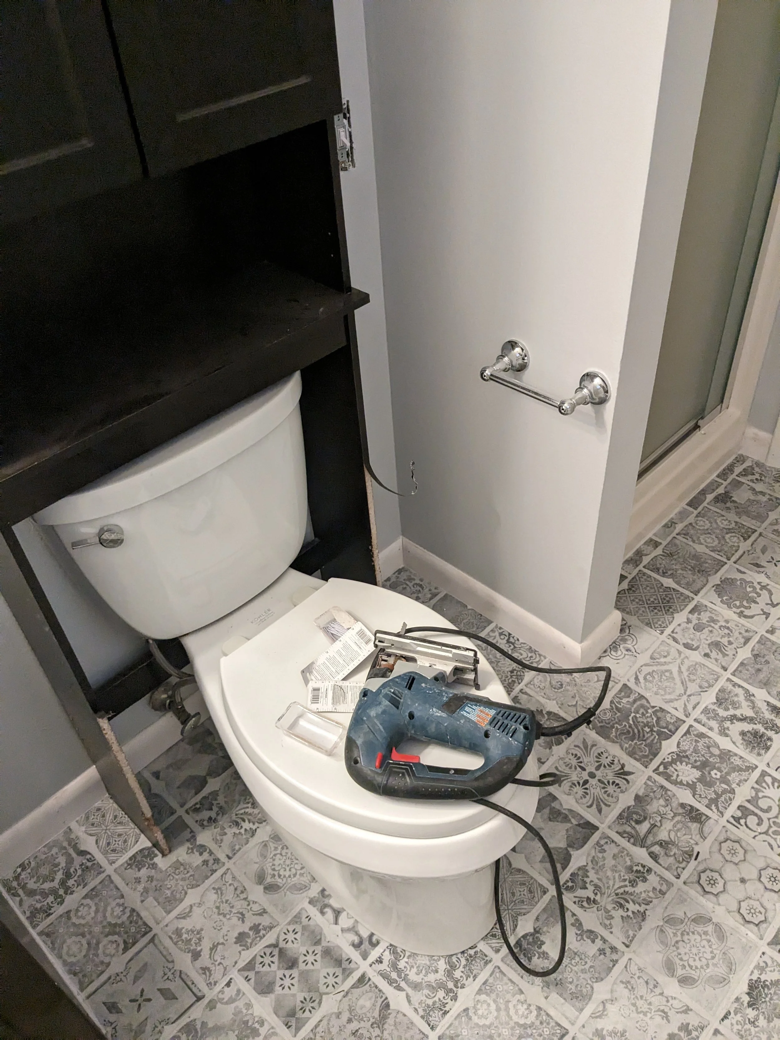 bathroom scene with jigsaw on toilet lid, black cabinet above, decorative tile floor