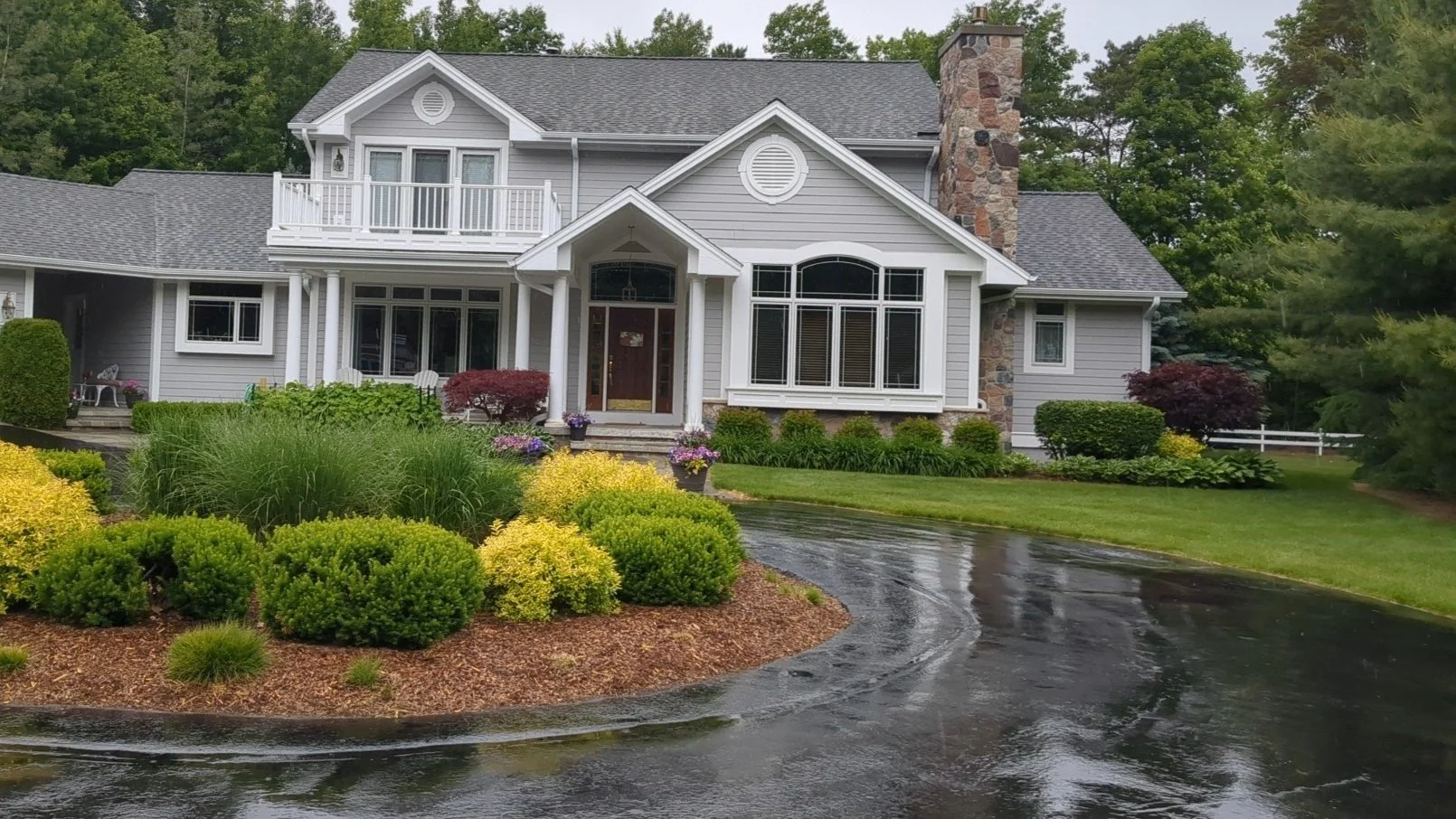 Beautiful suburban house with manicured landscape, grey siding, stone chimney, and large windows.