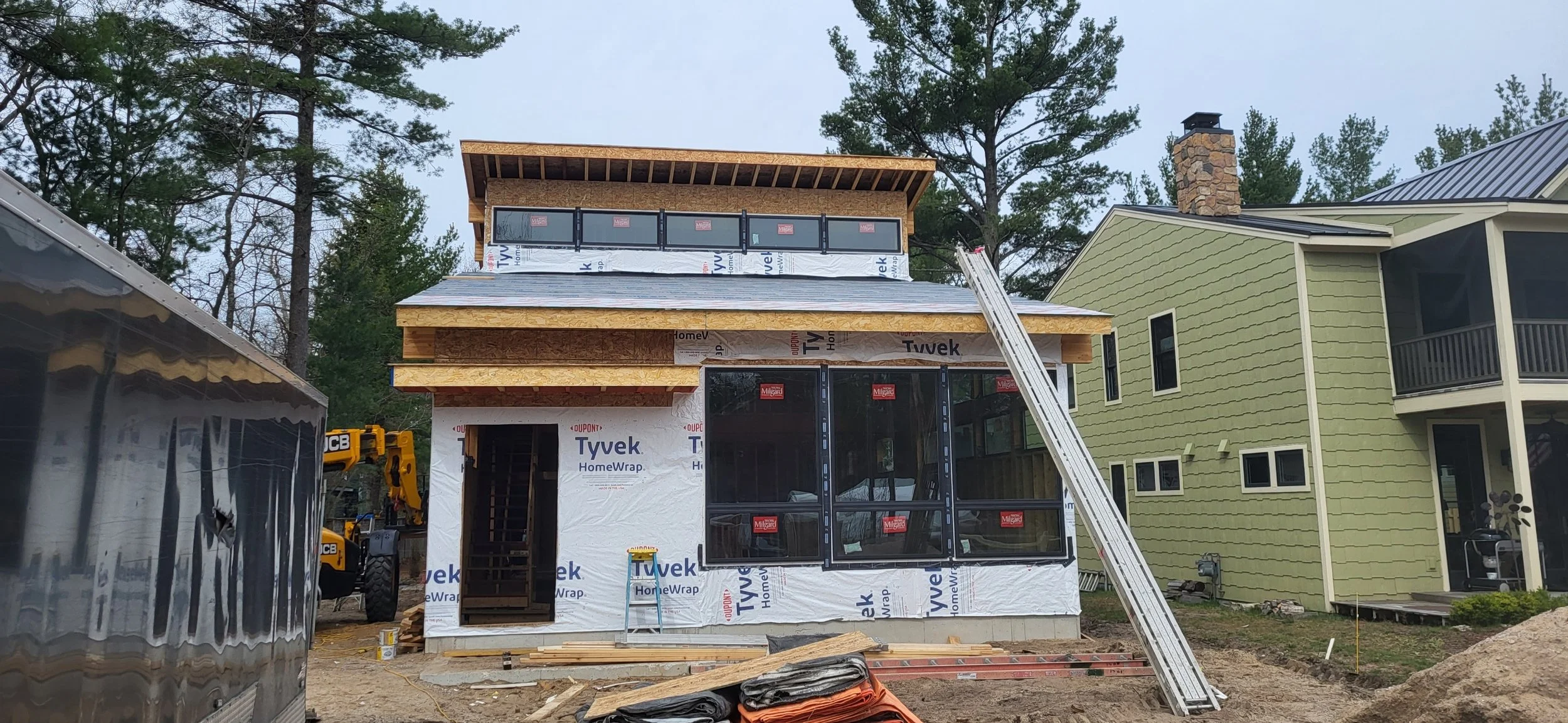 House under construction with Tyvek wrapping, large glass windows, and a wooden frame. A ladder is leaning against the structure. Nearby construction equipment and materials are visible.
