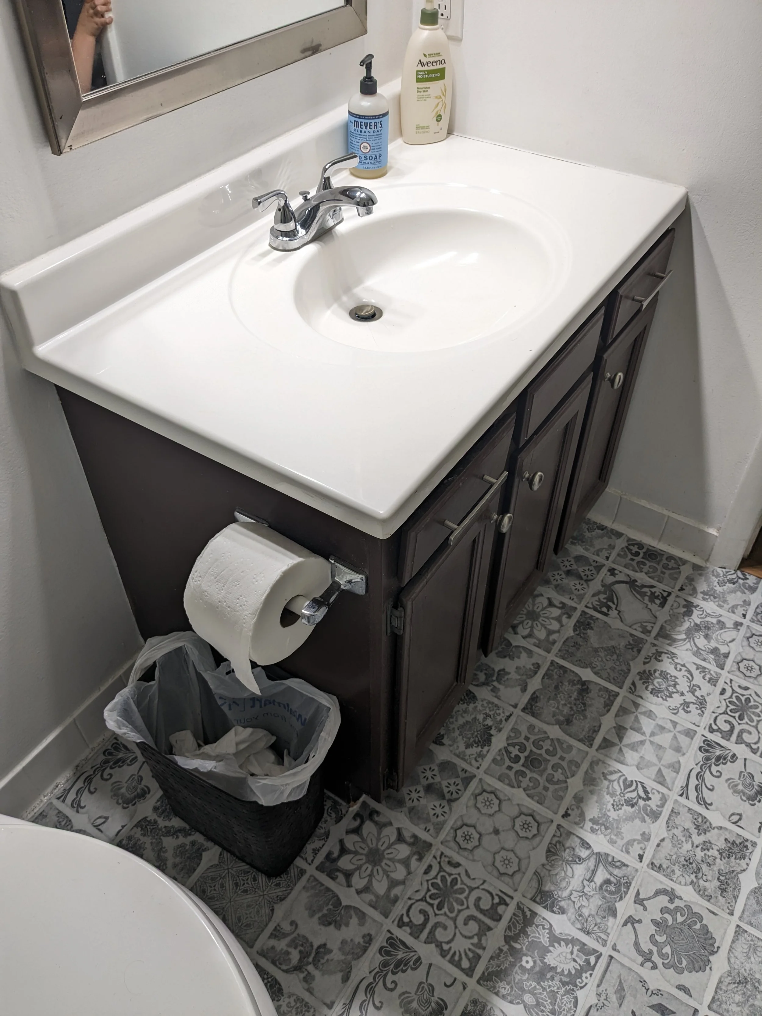Bathroom vanity with white sink, silver faucet, and two soap dispensers. Underneath is a dark cabinet with a toilet paper holder and a trash can. The floor has decorative tiles.