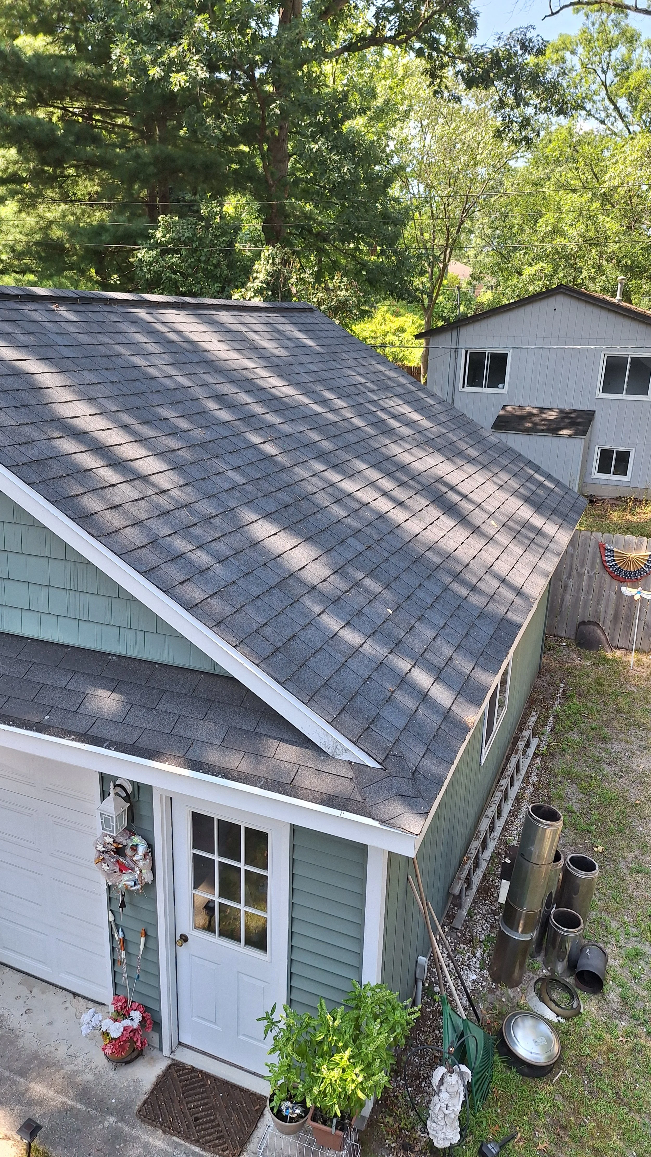 Aerial view of a green and white house with a shingled roof, surrounded by trees, showing a small garden with potted plants near the entrance.