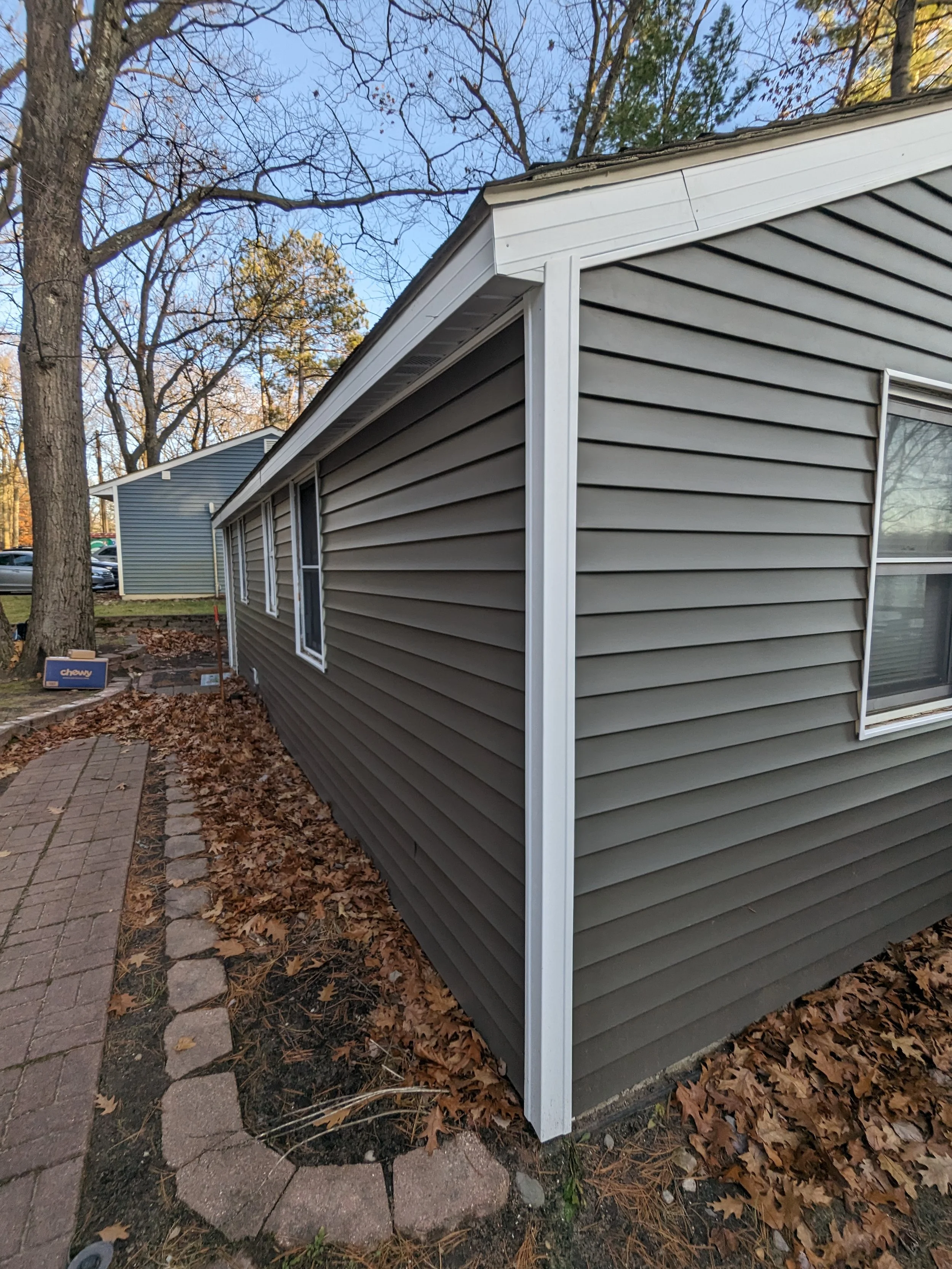 Exterior of a house with brown vinyl siding, white trim, and a window. Fallen leaves are scattered along a brick path and landscaped area. Trees and a clear sky are visible in the background.