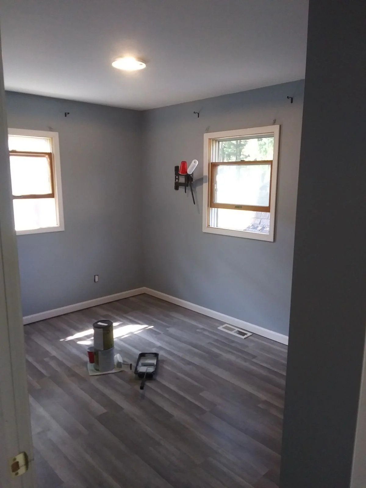 Empty room with gray walls, two windows, and wood flooring. Paint cans and a roller tray are on the floor.