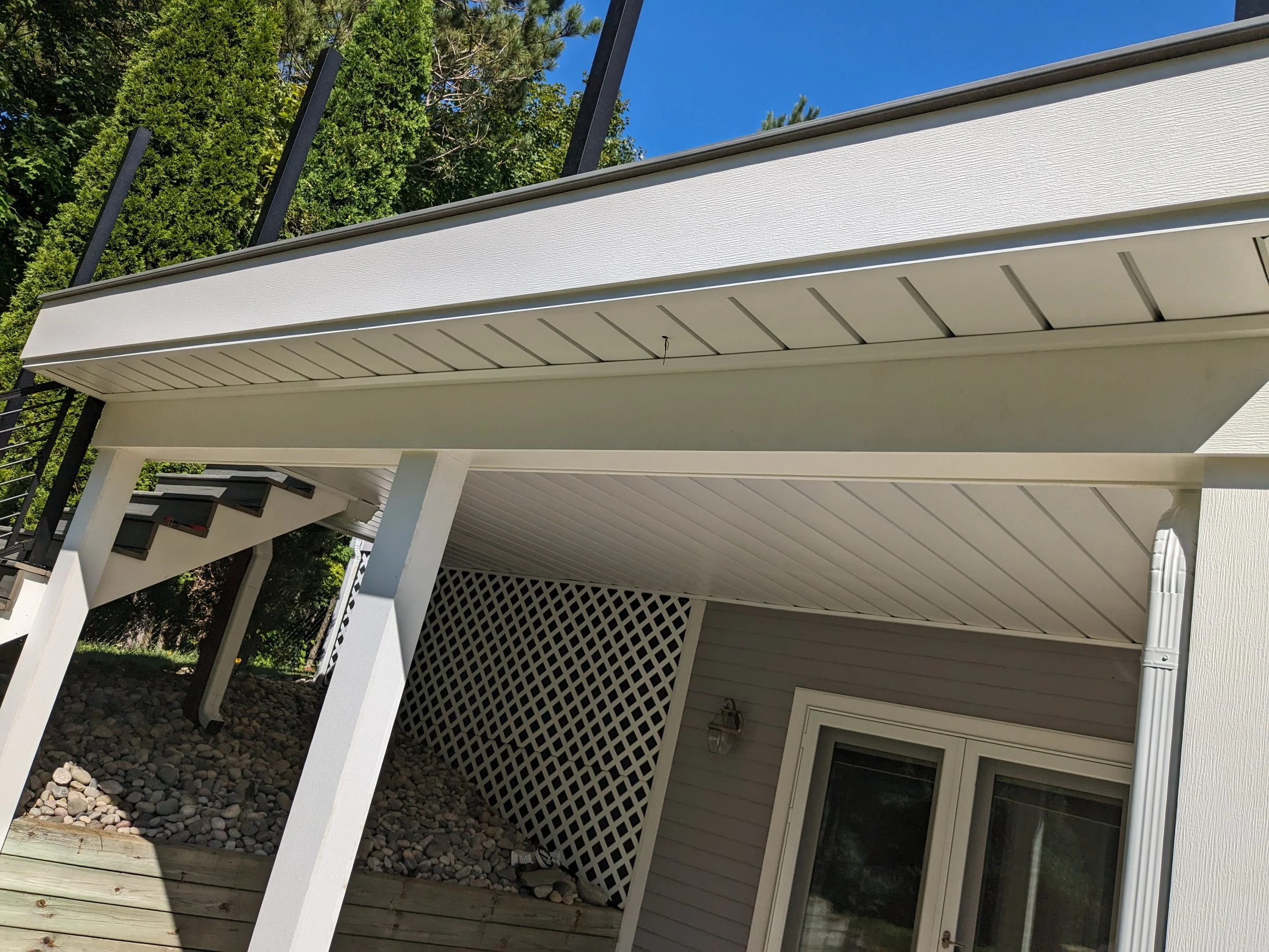 Underneath view of a house deck with white siding, support beams, lattice structure, and a staircase leading to the top of the deck. Surrounded by trees and clear blue sky.