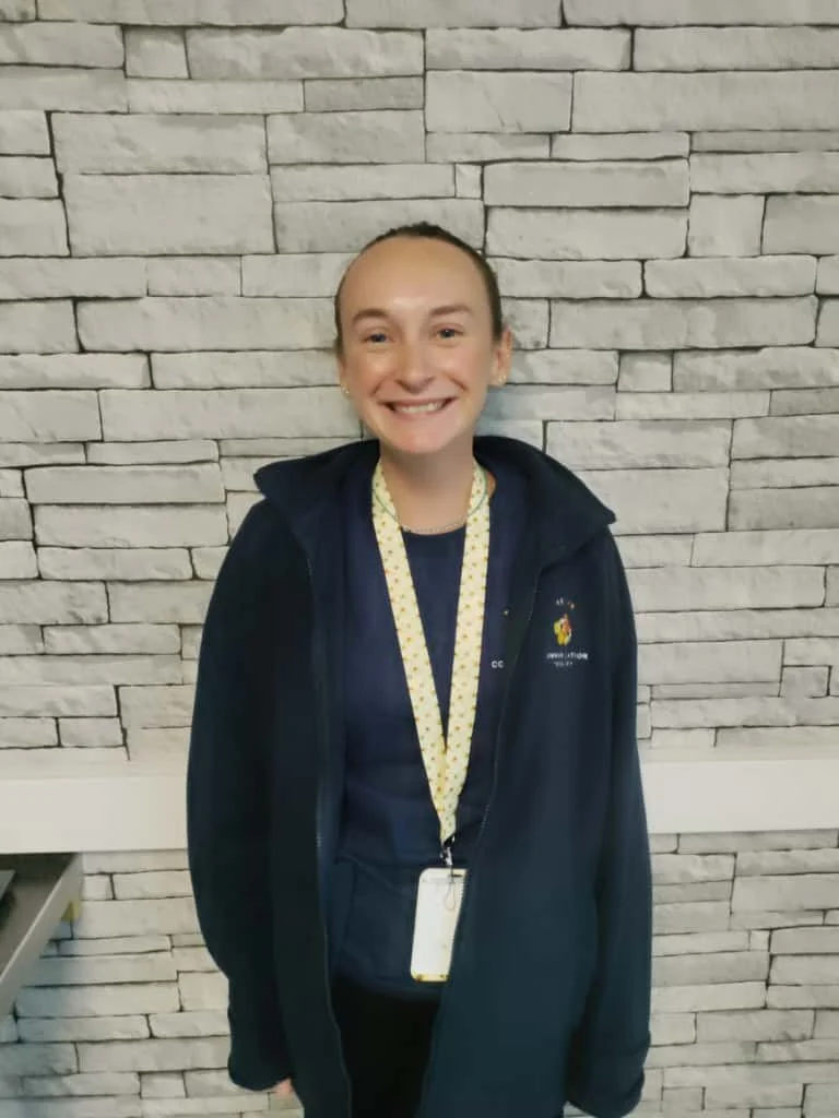 A young woman with light skin and a big smile standing in front of a white brick wall. She is wearing a dark jacket, a yellow lanyard with a badge, and a dark shirt underneath.