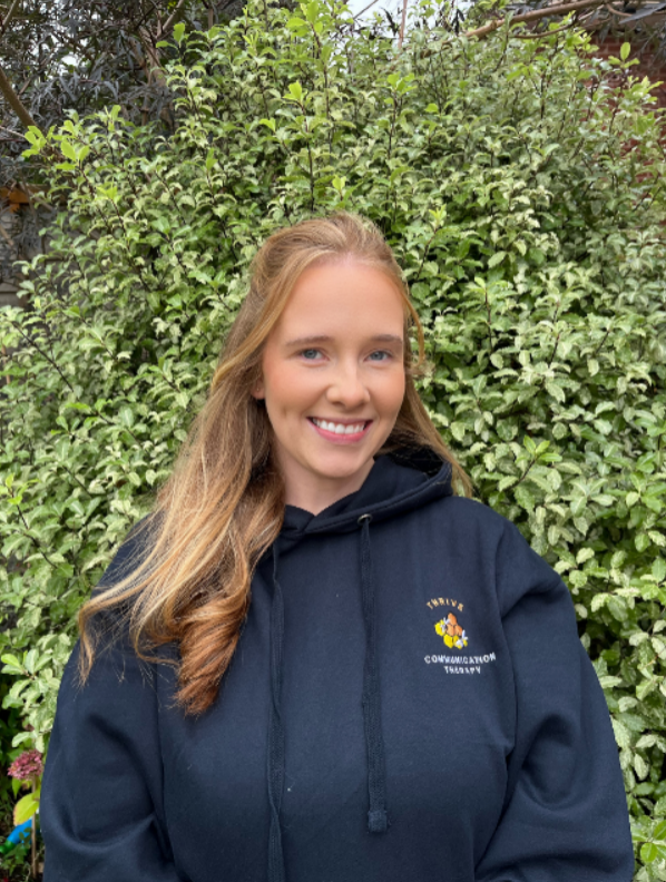 A young woman with long blonde hair smiling outdoors in front of a green leafy bush, wearing a navy hoodie with a logo that reads 'Thrive Communication Therapy'.