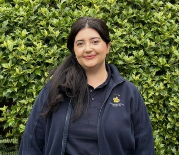 A woman with long dark hair smiling, standing in front of green leafy bushes, wearing a navy blue jacket and polo shirt with a logo.