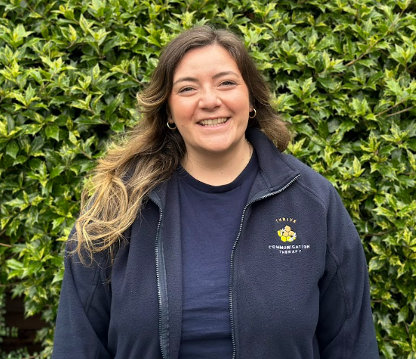 Woman with long, wavy hair smiling outdoors in front of green shrubbery, wearing a navy jacket with a logo that reads 'Thrive Communication Therapy'.