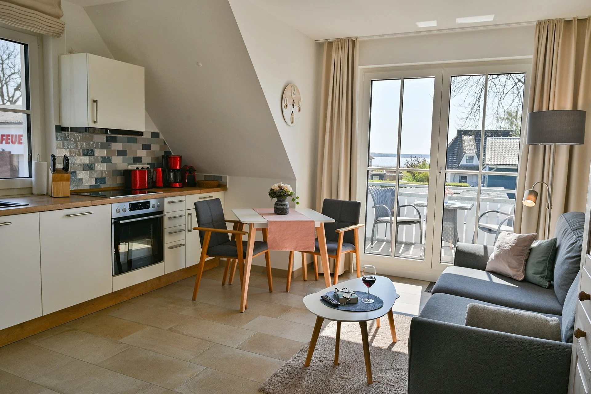 Wohnzimmer mit Küche, Esstisch und Balkon, Blick aufs Wasser, Sonnenlicht, gemütliche Einrichtung, Sofa, Weinglas, Fernseher, Vorhänge.