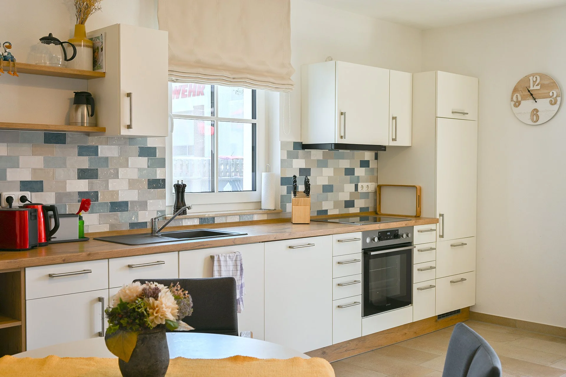 Moderne Küchenzeile mit weißen Schränken und Holzarbeitsplatte, darunter ein Herd und ein Spülbecken, im Hintergrund eine Fenster mit beige Vorhang, auf dem Tisch im Vordergrund eine Vase mit Blumen.