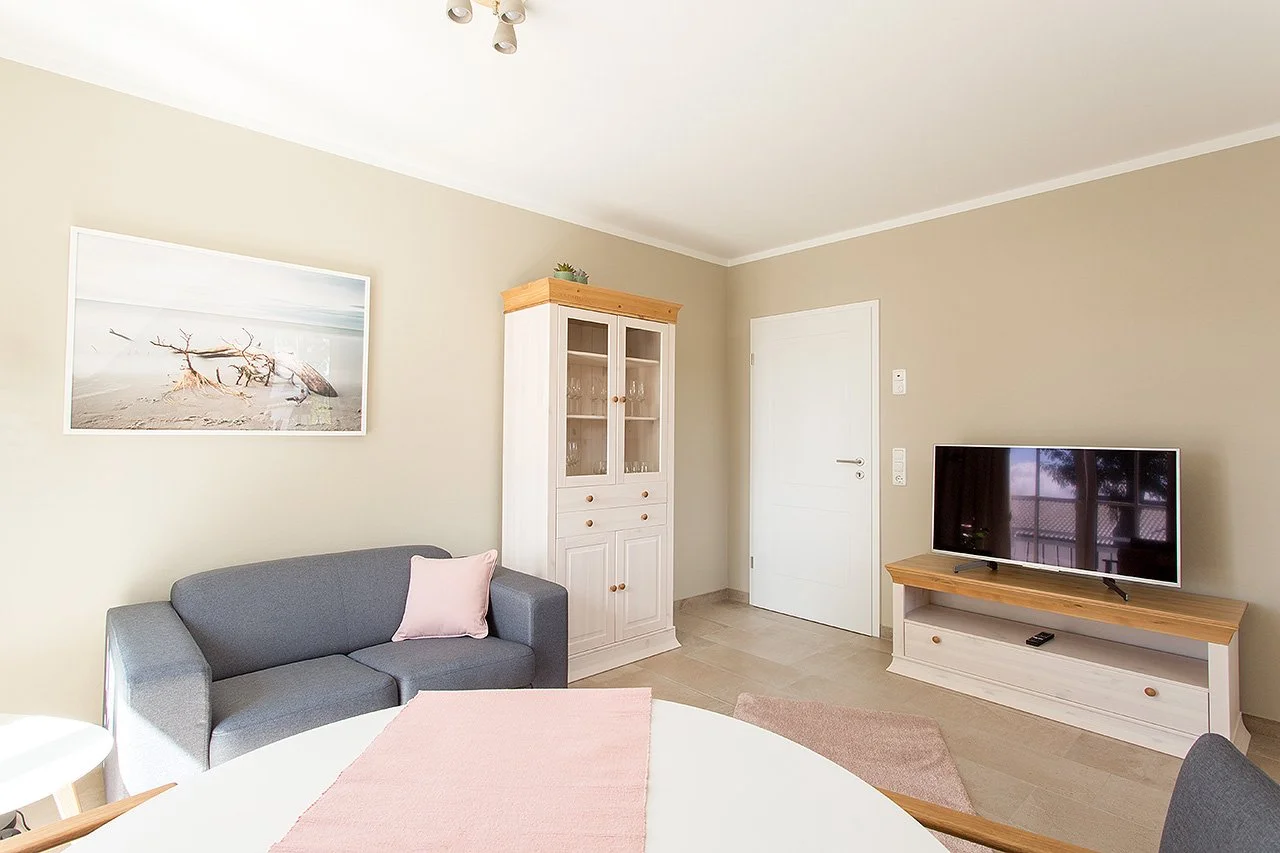 Moderner Wohnraum mit grauer Couch, weißem Wandregal, Fernseher, Wandbild eines Strands mit Treibholz, beige Wände, Holzmöbel, Türen und Vorhänge.