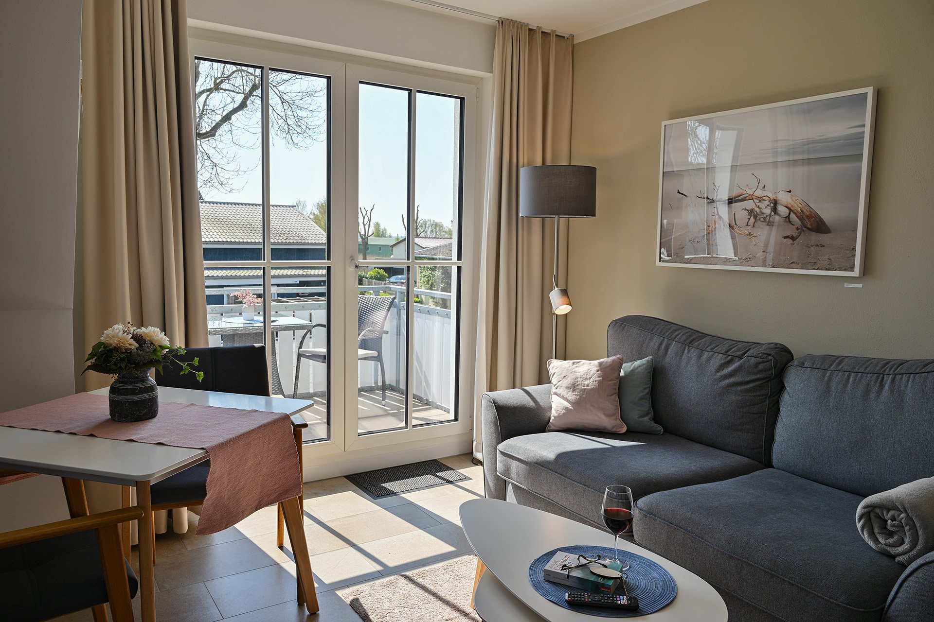 Gemütliches Wohnzimmer mit Balkon, grau Sofas, einem kleinen Tisch mit Wein und Fernbedienung, einem Esstisch mit Blumenvase, großen Fenstern und Sonnenlicht.