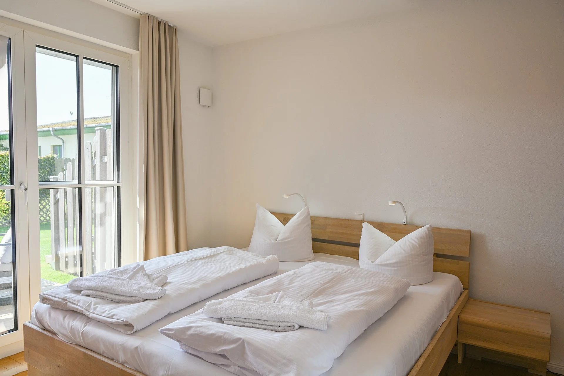 Ein modernes Hotelzimmer mit Doppelbett, weißen Bettwäsche, Kopfkissen, Nachttischen und Leselampen, große Fenster mit Vorhängen, Blick nach draußen auf Grünflächen.