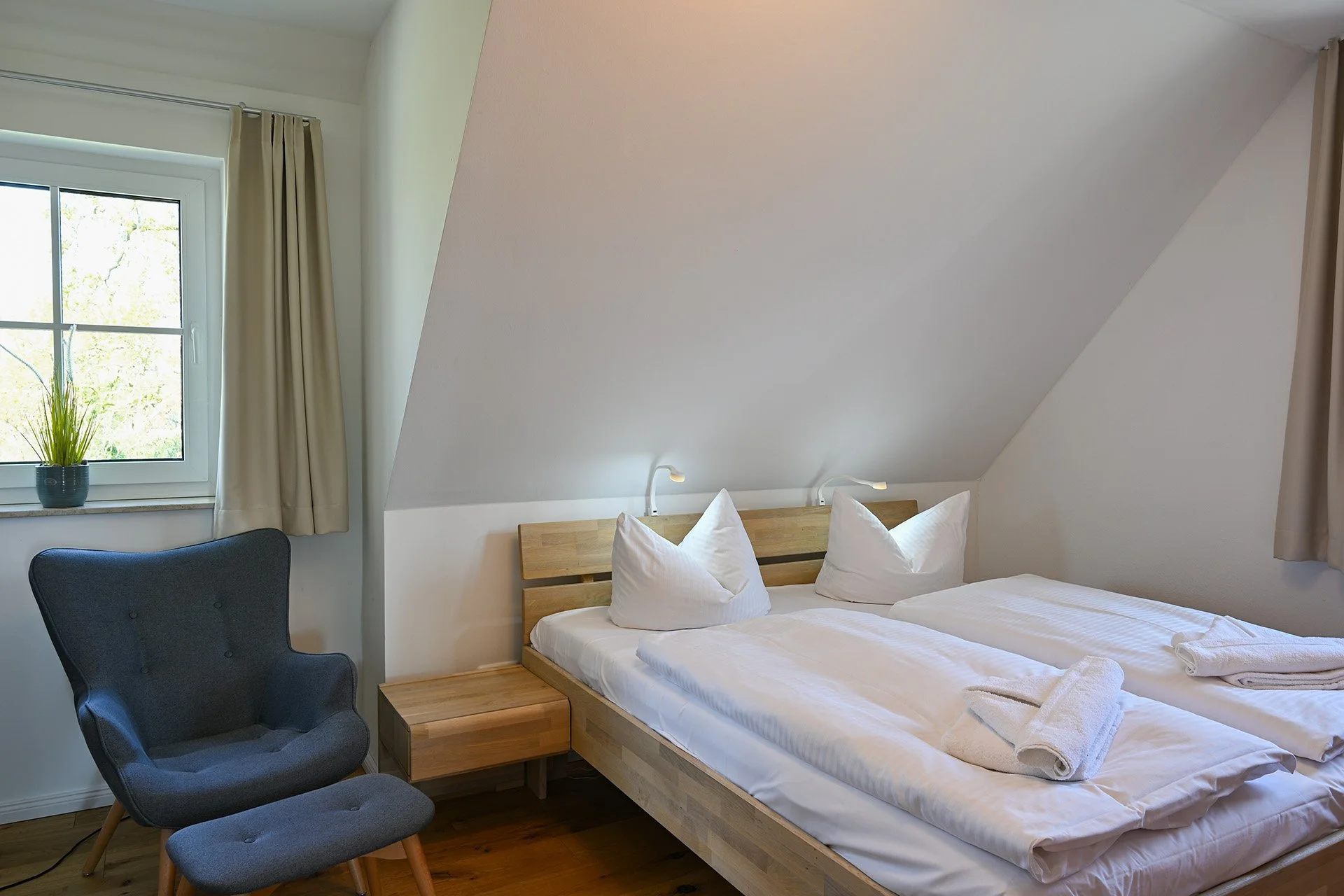 Ein Schlafzimmer mit einem Doppelbett, weißen Bettwäsche und Kissen, ein Nachttisch und ein bequemer Sessel neben einem Fenster mit beige Vorhängen.