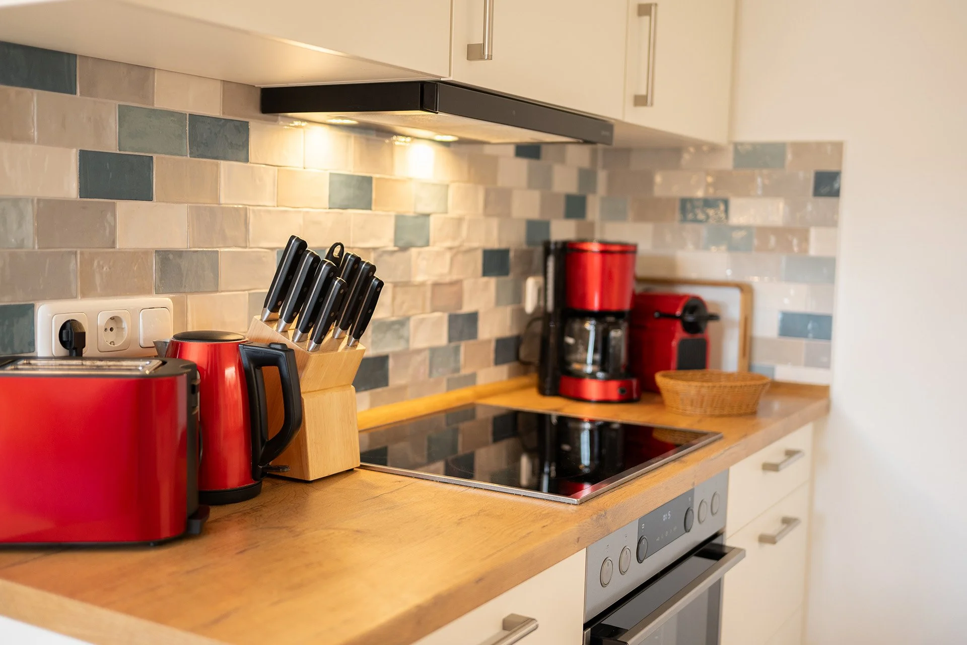 Küche mit Holzarbeitsplatte, beige Fliesen mit blauen Akzenten, rotem Kaffeeautomaten, Toaster, Messerblock, Wasserkocher, weitere Küchengeräte