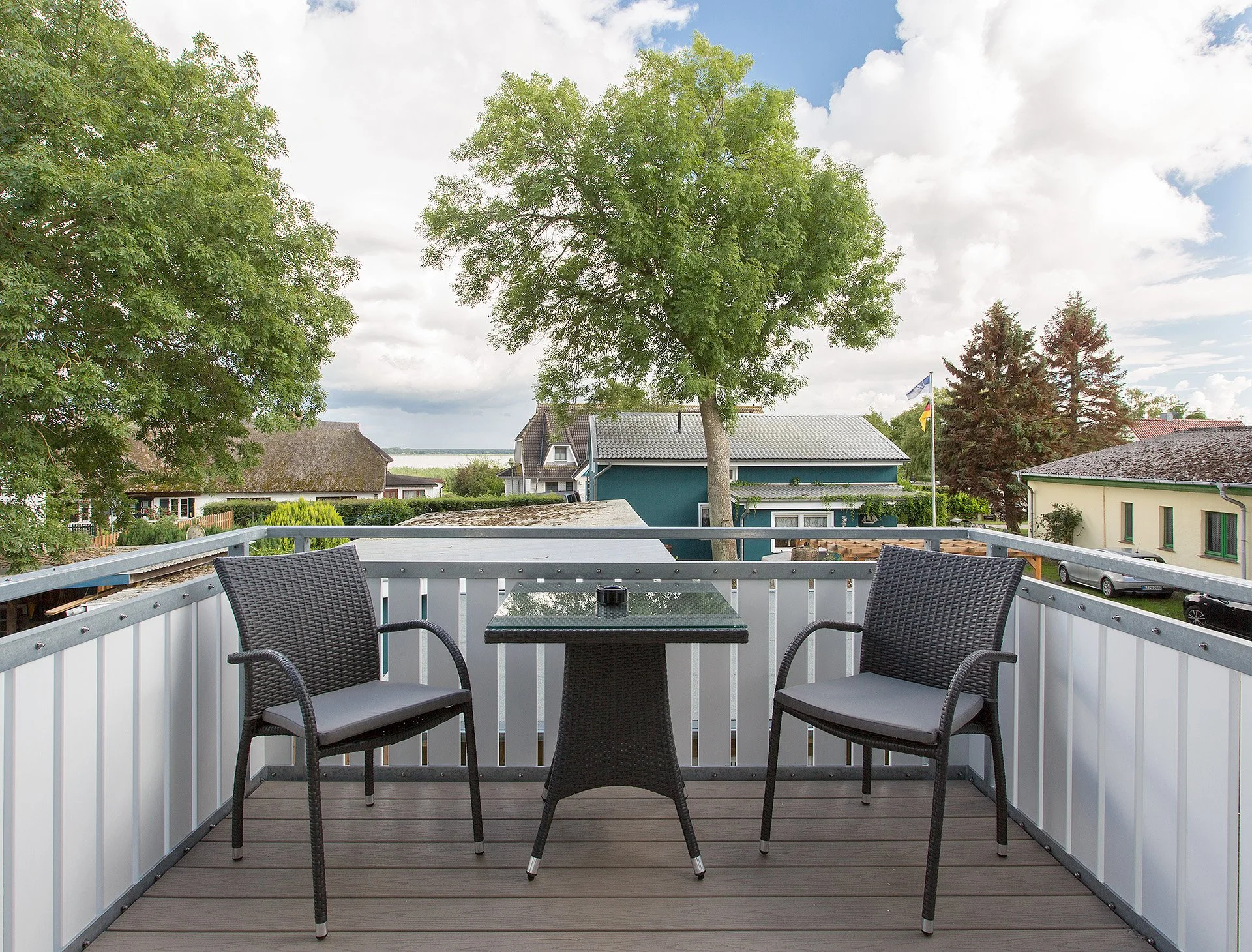 Balkon mit zwei Stühlen, einer kleinen Tisch in einer ruhigen Nachbarschaft mit Bäumen und Häusern, Flaggen im Hintergrund, bewölkter Himmel.