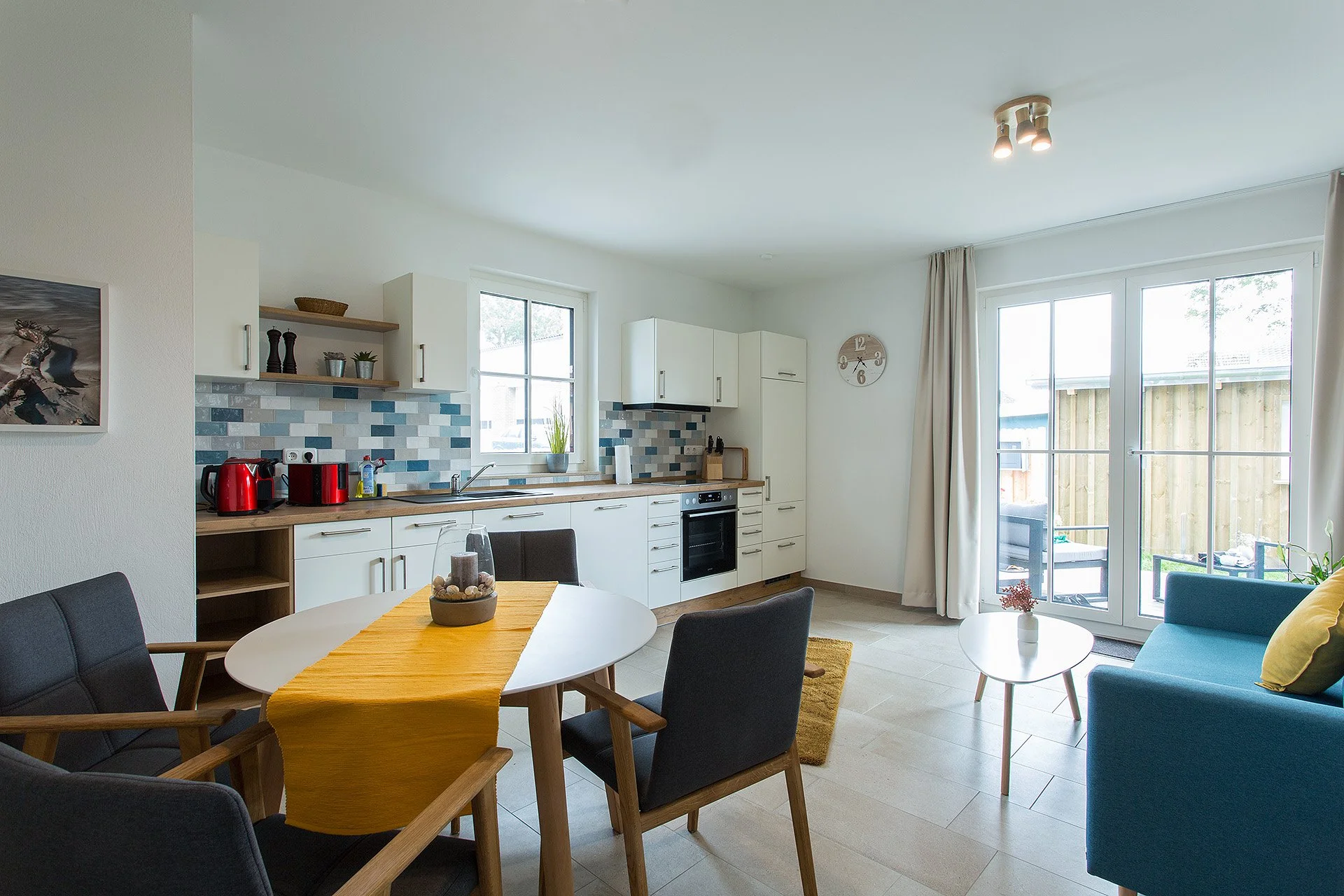 Moderne offene Küche und Wohnzimmer mit Essbereich, große Fenster und Gartentür, gemütliche Sitzmöbel, bunte Dekorationen.