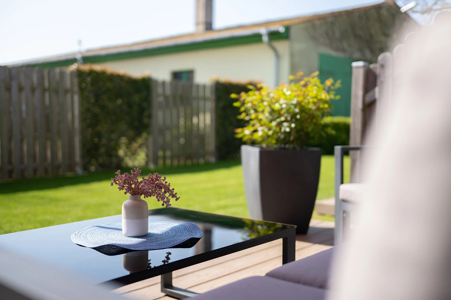 Gedeckter Sitzbereich mit einer schwarzen Glastisch, weißem Blumentopf mit rosa Blumen, auf einer weißen Unterlage, im Garten mit grünem Rasen, Holzzaun und Haus im Hintergrund, sonnig und hell.