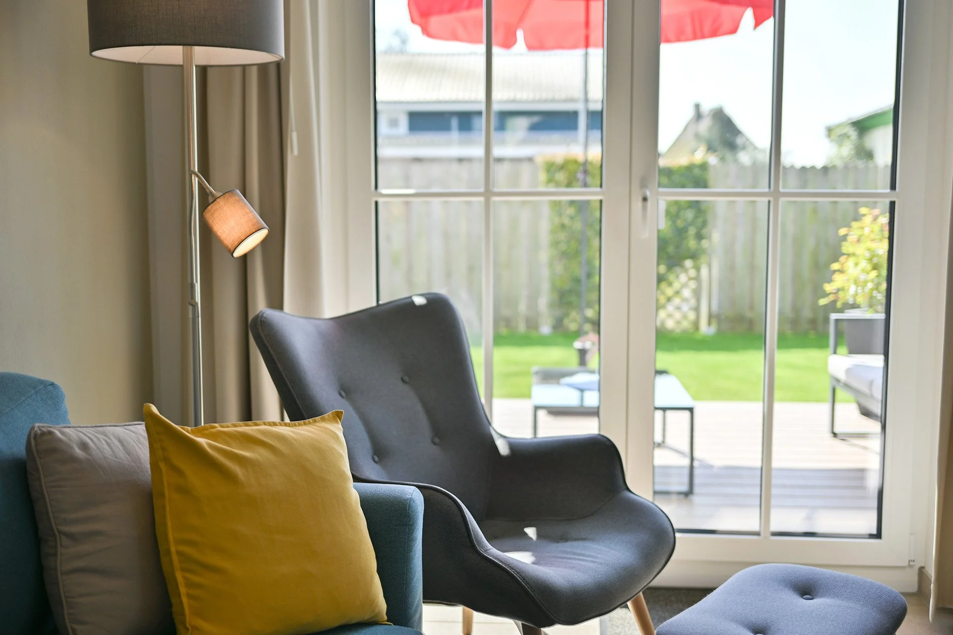 Kombination aus dunklem Sessel, blauen und gelben Kissen an einem Sofa, Stehlampe, große Glasschiebetür mit Garten und Terrasse im Hintergrund bei Tageslicht.