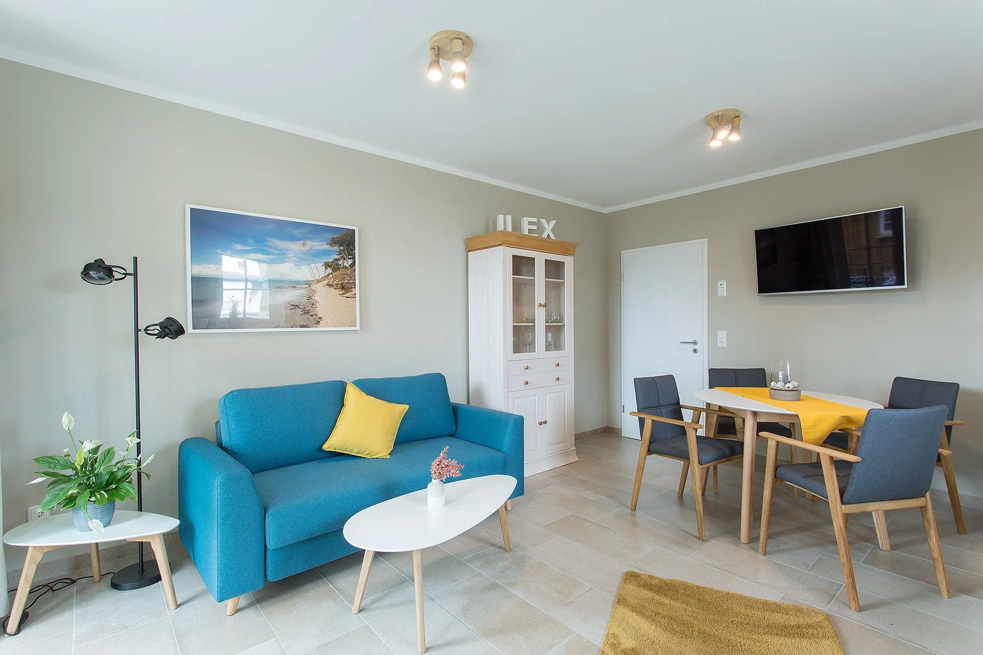 Geräumiges Wohnzimmer mit blauer Couch, einem weißen Couchtisch, einer Tischlampe, einer grünen Pflanze, Wandbild eines Strandpanoramas, einem weißen Holzschrank, einem Esstisch mit gelbem Tuch und vier Stühlen sowie einer Wandtafel und einem Fernseh