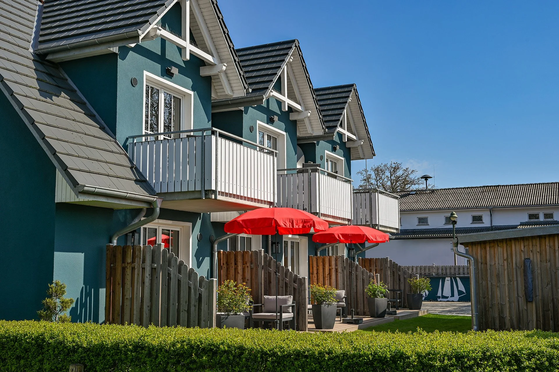 Mehrparteienhaus mit blauer Fassade, weißen Balkonen, grauen Dachziegeln und einem Garten mit roten Sonnenschirmen und Pflanzgefäßen.