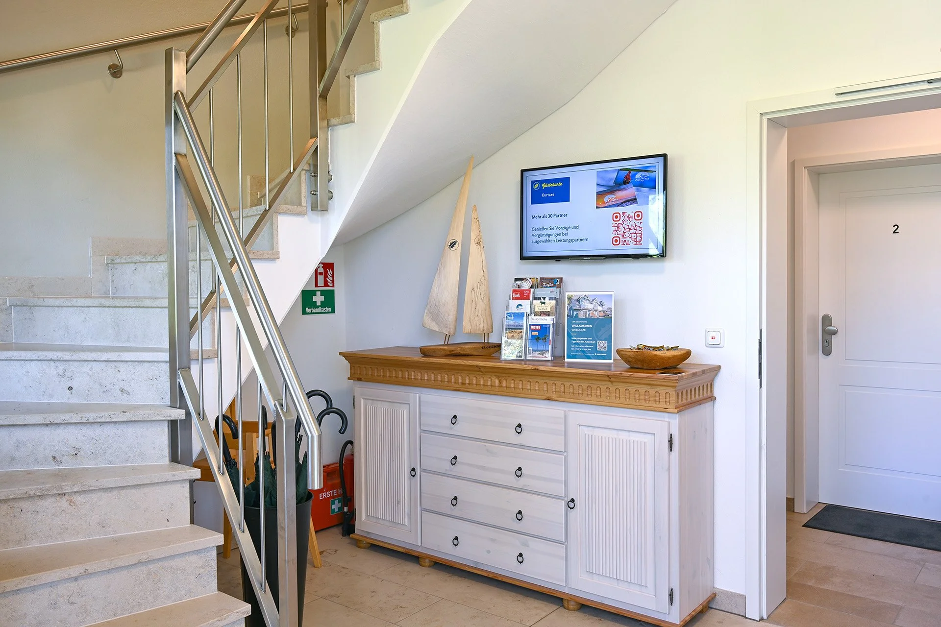Flur mit Treppe, weißem Sideboard, Fernseher an der Wand, Revisionskasten, Schlüsseln auf dem Sideboard, Briefkarten, Schüssel, hinter Türen mit Nummer 2