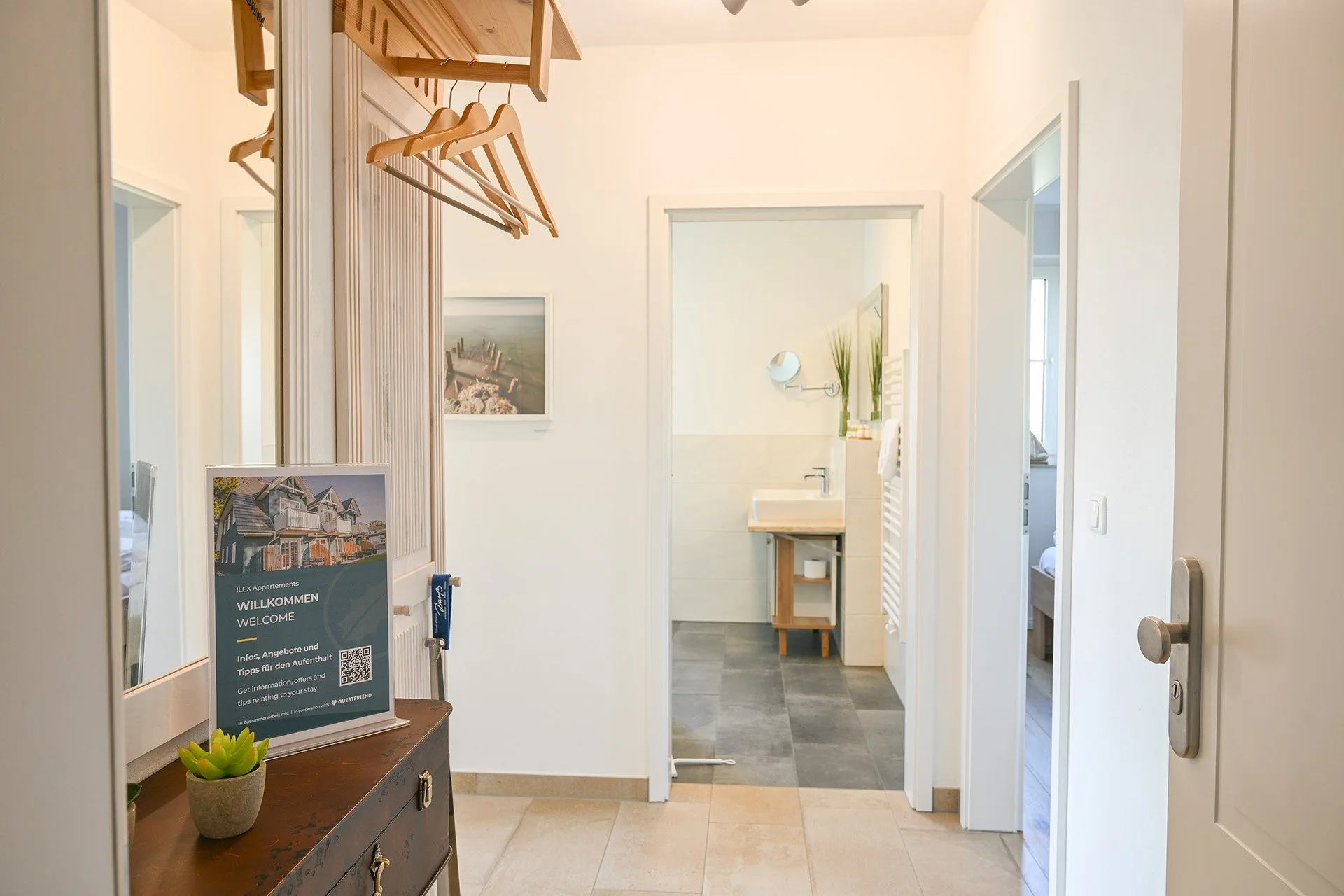 Eingangsbereich einer Ferienwohnung mit Willkommensschild, Spiegel, Pflanzen und Kleiderhaken, Blick in das Badezimmer mit Waschbecken und Fliesenboden.