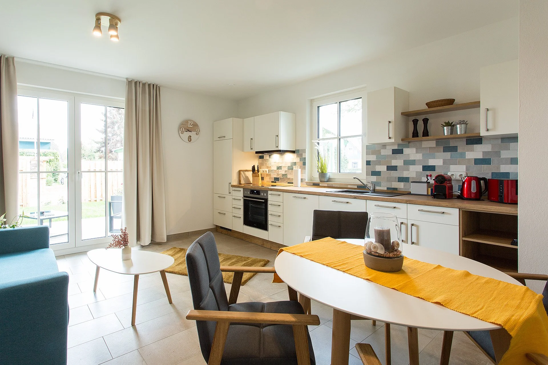 Moderner Küchen- und Wohnbereich mit Fenster, Esstisch, Stühlen, Küchenzeile, Backofen, Herd, Geschirrspüler, Geschirr, Pflanzen, Wanduhr, und Terrasse im Hintergrund.