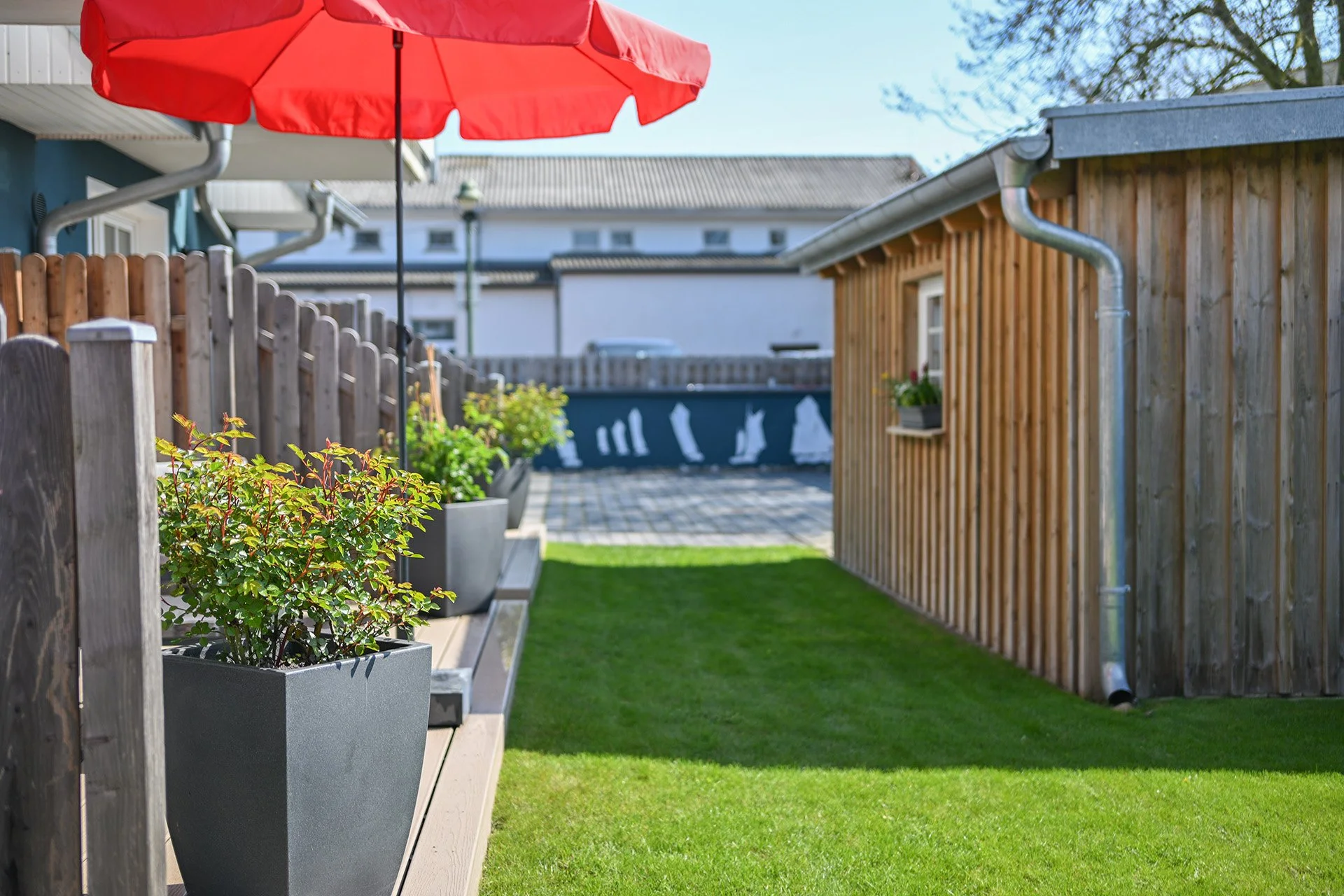 Ein Garten mit grünem Rasen, Holzzäunen, großen Pflanzkübeln mit Büschen, einer roten Schirm und einem Holzhaus.