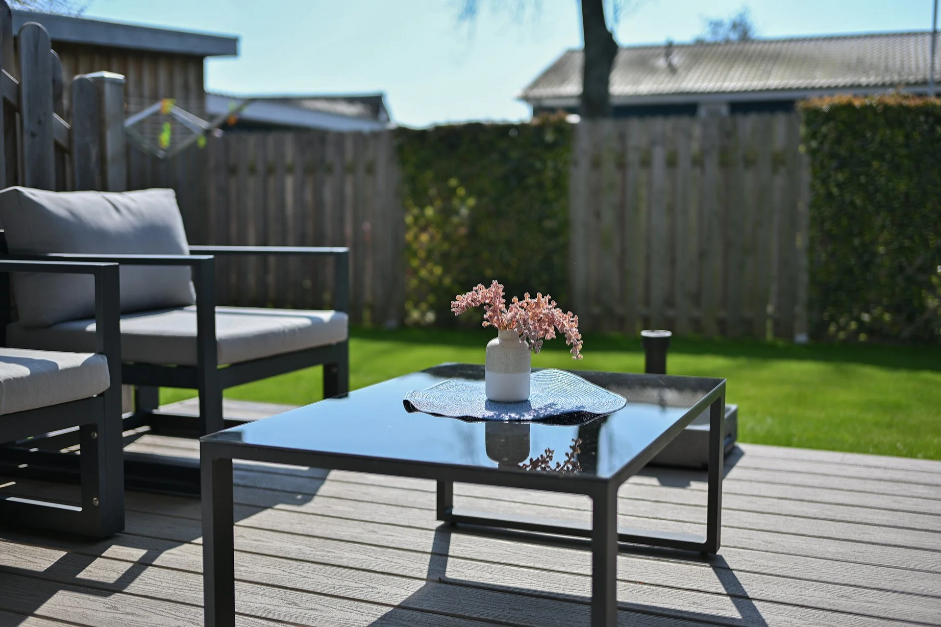 Garten mit Sitzmöbeln, einem schwarzen Couchtisch mit Vase und Blumen, grünem Rasen, Holzzaun und Häuser im Hintergrund bei sonnigem Wetter.