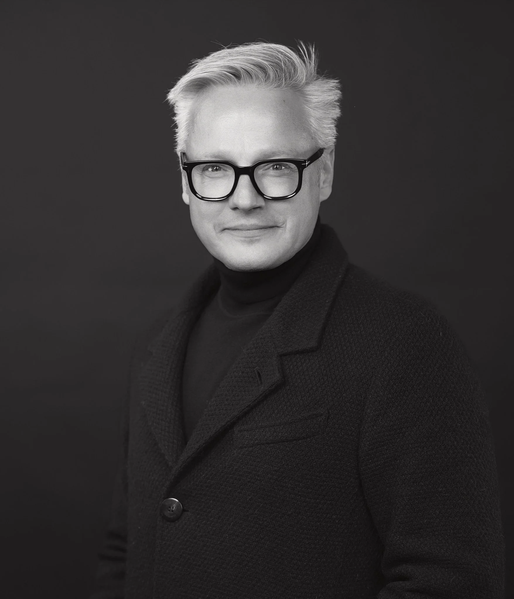 Schwarz-Weiß-Bild eines Mannes mit grauem Haar, Brille und dunklem Pullover, lächelnd, vor dunklem Hintergrund.