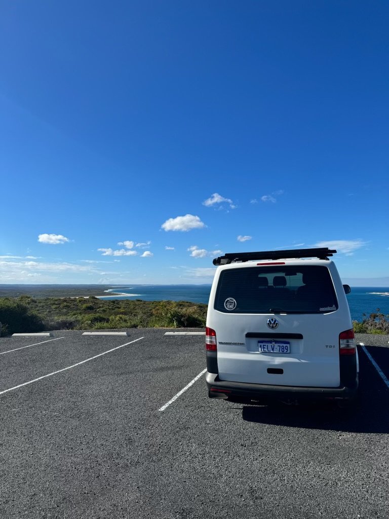 White Volkswagen van parked in an empty parking lot near the coast, overlooking the ocean and a clear blue sky with scattered clouds.