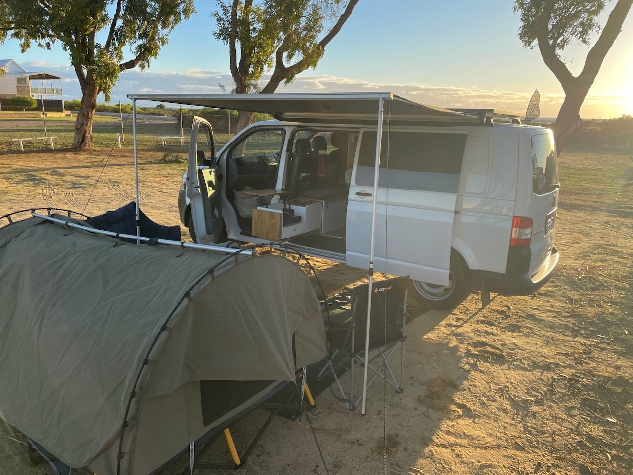 A white camper van with an extended awning is parked on a sandy area, with a green tent, two folding chairs, and a small table set up in front of it, under a tree during sunset.