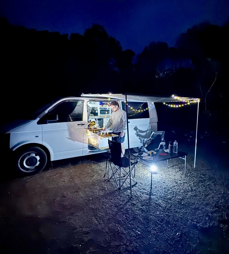 A man cooking food at night outside a white van decorated with string lights, set up with camping chairs and tables on a dirt surface in a wooded area.