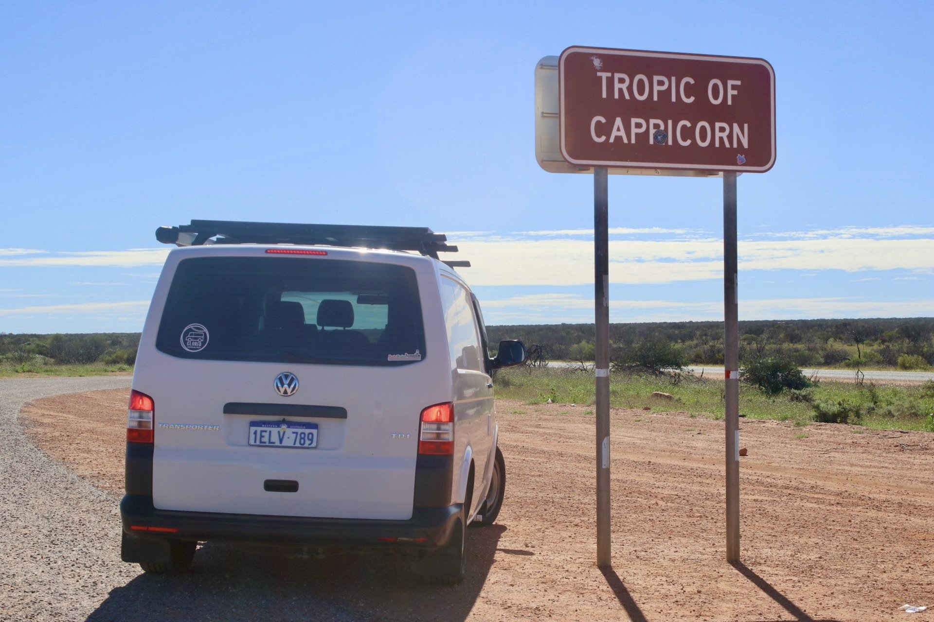 A white Volkswagen Transporter van parked on a dirt road next to a sign that reads "Tropic of Capricorn" in a flat, open landscape with blue sky and scattered clouds.