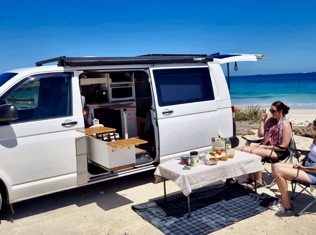 A white camper van parked on a sandy beach with a table set for breakfast, including fruits and juices, and a woman sitting in a folding chair near the ocean, waving.