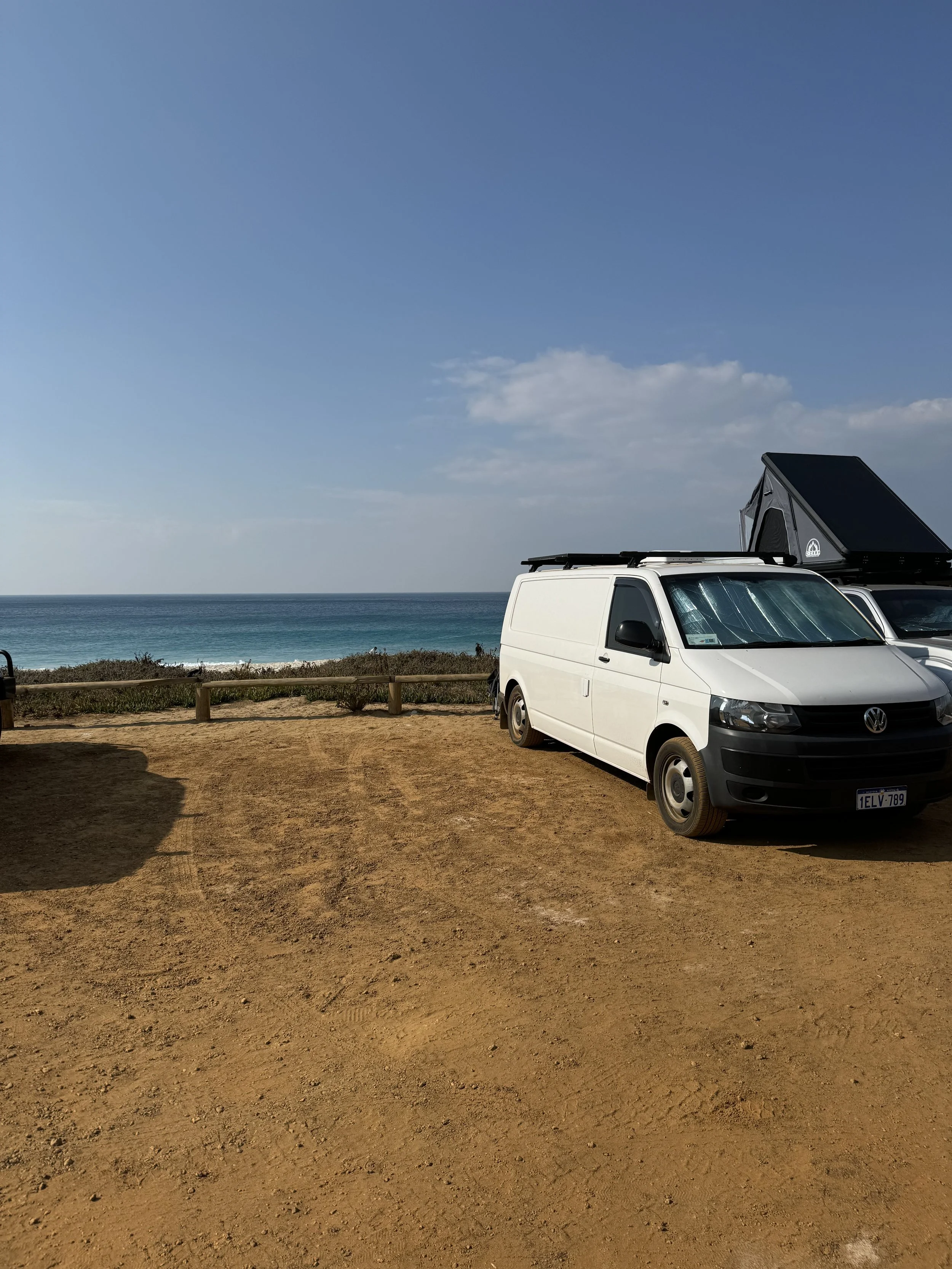 White van parked on sandy area near the ocean with clear blue sky and some clouds.