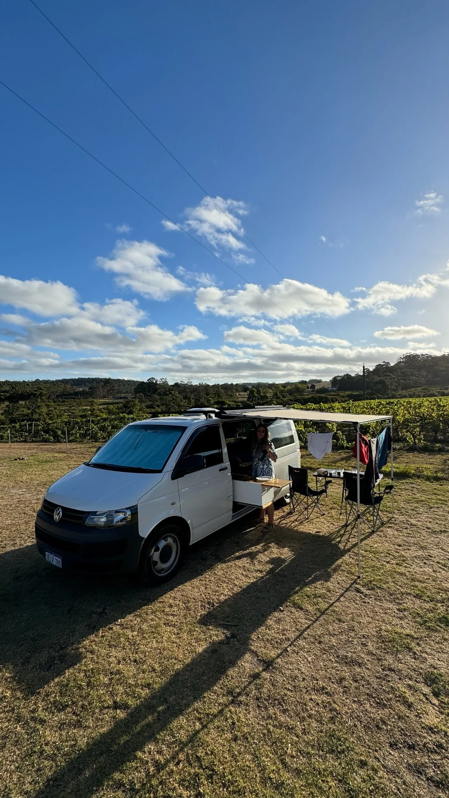 A woman standing inside a camper van with a canopy set up in an open field surrounded by greenery and distant trees, with a blue sky and white clouds overhead.