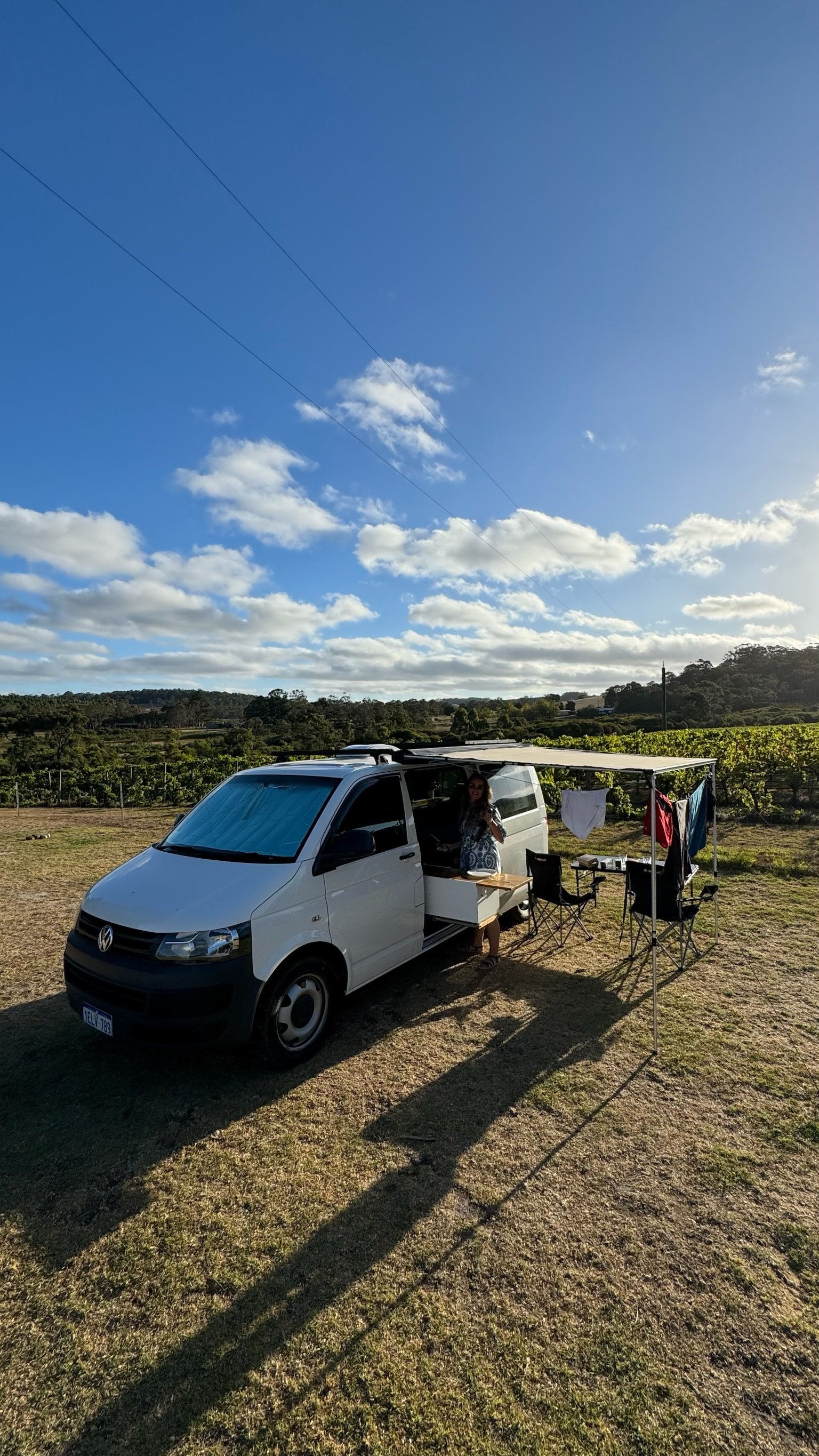 A woman preparing food at a mobile kitchen setup outside a white Volkswagen van with an extended awning in a grassy field on a sunny day with scattered clouds.