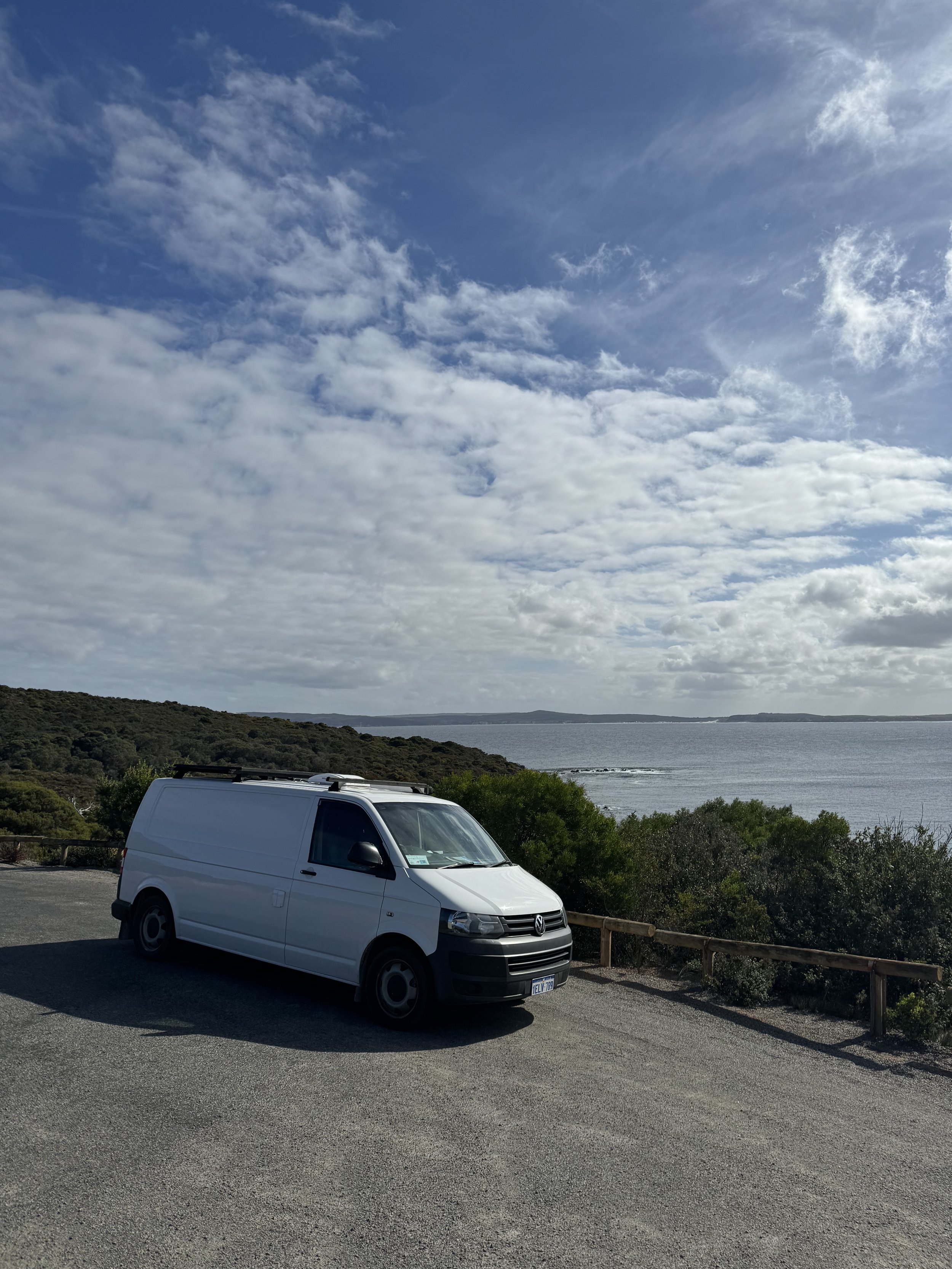 A white Volkswagen van parked on a gravel lot near the coast, with a view of the ocean, rolling hills, and partly cloudy sky in the background.
