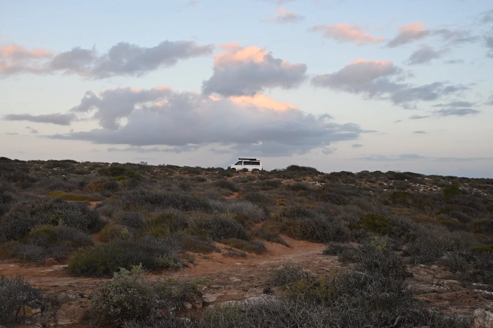 A white van with a solar panel on top parked on a hill in a desert landscape with low shrubs and clouds in the sky.