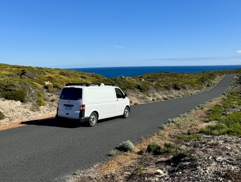 White van parked on a winding coastal road with ocean and blue sky in the background.