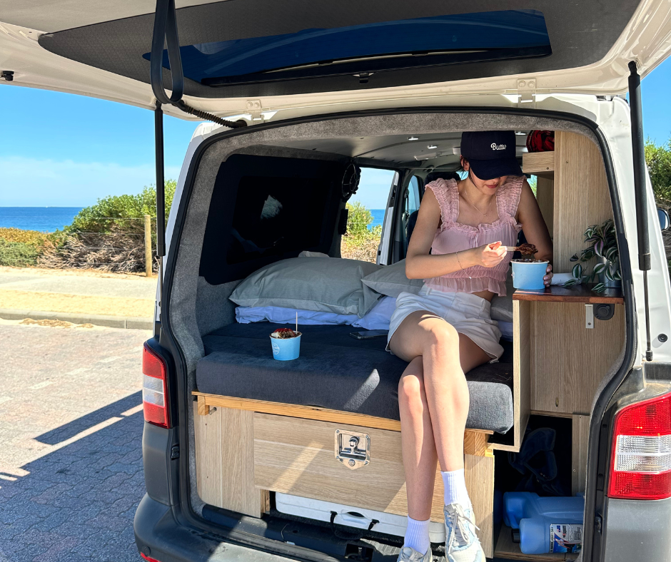A woman sitting inside the back of a camper van by the beach, eating ice cream. The van has a bed, pillows, and wooden shelves, with a scenic ocean view in the background.