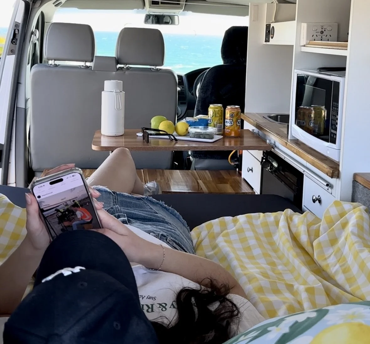 Inside a camper van with a view of the ocean, a person is lying on a bed using a smartphone, with a yellow and white checkered blanket. The van has a small kitchen area with a microwave, a sink, and snacks and drinks on a table, including lemons and cans of sparkling water. The front seats and windshield are visible, showing the ocean outside.