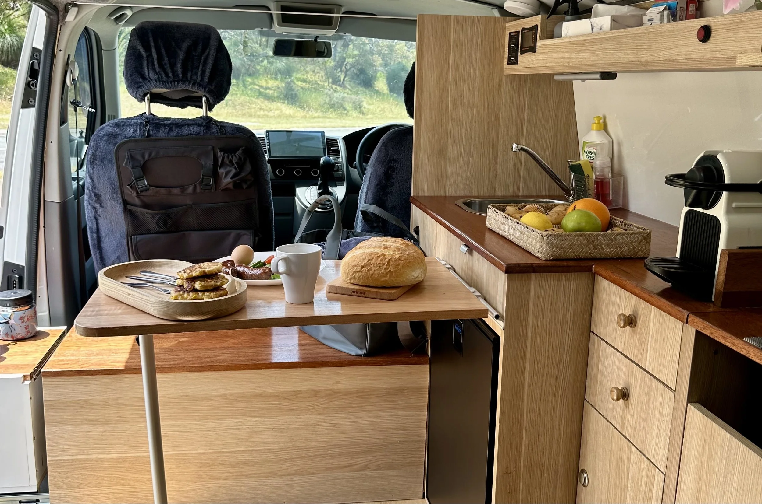 Interior of a camper van, showing a small kitchen area with a wooden countertop, sink, and a few appliances. There is a table with breakfast items including a loaf of bread, a plate of pancakes, a cooked sausage, an egg, a mug, and some fruit on it. The driver's and passenger's seats are visible at the front, with a view of greenery outside the windshield.