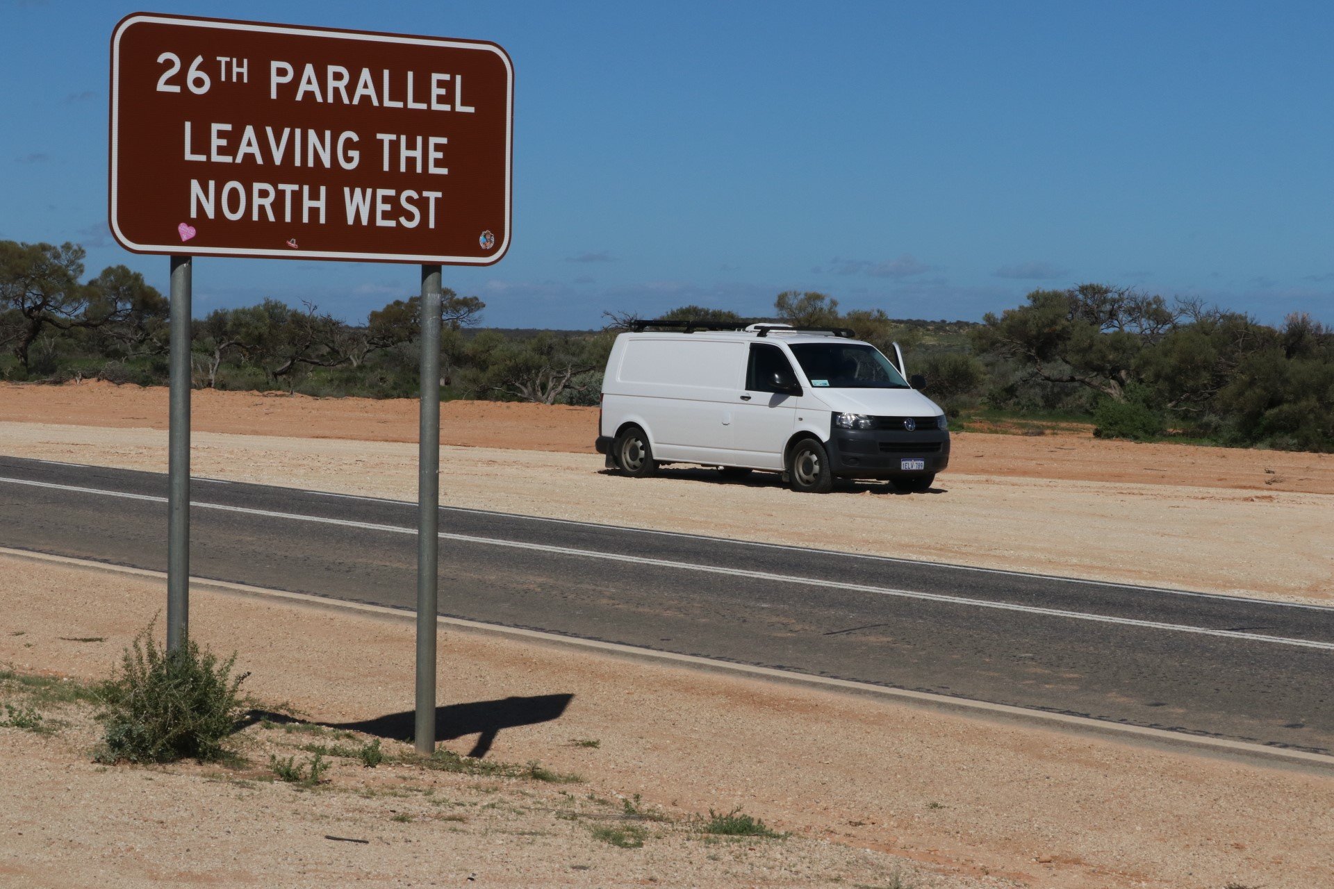 A brown road sign indicating '26th Parallel Leaving the North West' is posted off the side of a rural highway in a desert landscape. A white van is parked on the sandy shoulder nearby, with sparse bushes and trees under a clear blue sky in the backgr